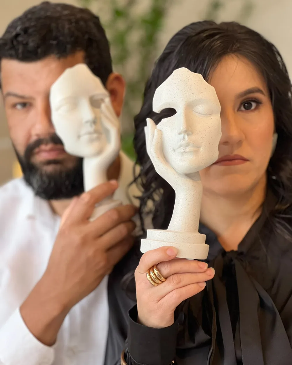 Luiz Paulo e Alê segurando esculturas de rostos diante dos seus próprios rostos